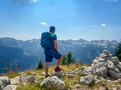 Hiker with backpack standing on rocky summit in the Julian Alps, overlooking mountain ranges under blue sky with stone cairn nearby.