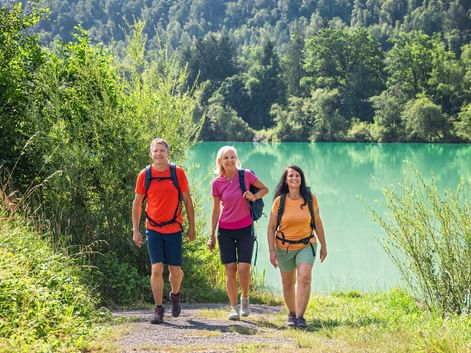 Three hikers with backpacks walking on a path along the turquoise Danube River. Dense green forest covers the hillside in the background.