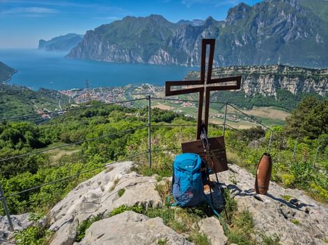 Wooden summit cross with blue backpack on rocky peak above Lake Garda. View shows Torbole-Nago, mountains, and northern lakeshore.