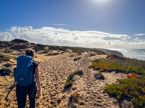 Hiker with blue backpack walking on sandy coastal path at Praia do Brejo Largo, Portugal. Ocean and dramatic clouds visible under sunny sky.