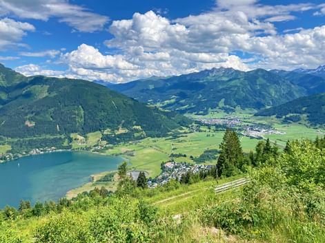 Panoramablick von der Schmittenhöhe über den Zeller See, grünes Tal und Bergketten im Pinzgau unter blauem Himmel mit weißen Wolken.