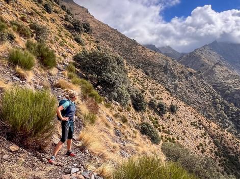 Wanderin mit Rucksack auf schmalem Bergpfad in den Alpujarras, Andalusien. Steile felsige Hänge mit Grasbüscheln und dramatische Berggipfel unter bewölktem Himmel.