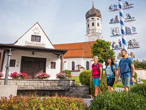 Three hikers with backpacks walk past a traditional Bavarian smithy and white church with onion dome in Andechs. A decorated maypole stands nearby.
