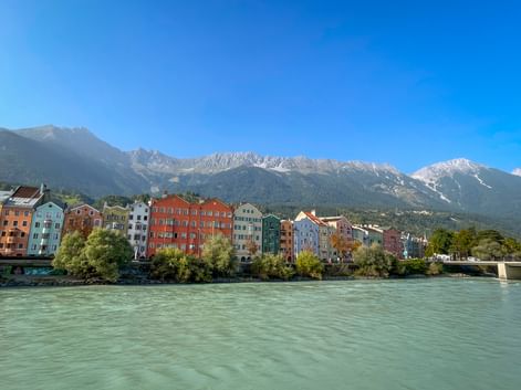 Row of colorful historic houses along the Inn River in Innsbruck with snow-capped Alpine mountains in the background under clear blue sky.