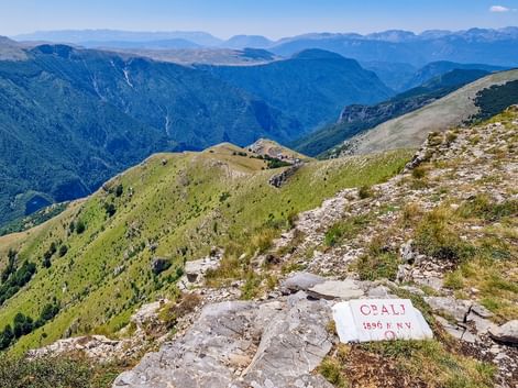 White summit marker reading 'OBALJ 1896 m n.v.' on rocky mountain peak with panoramic view of Bjelasnica mountain ranges in Bosnia.