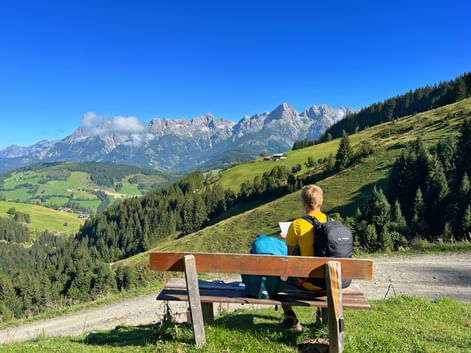 Two hikers with backpacks sit on a wooden bench overlooking green alpine meadows and the rocky peaks of the Kitzbühel Alps under a blue sky.