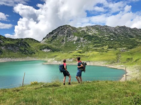 Zwei Wanderer mit Rucksäcken bewundern den türkisfarbenen Formarinsee, umgeben von grünen Almwiesen und felsigen Bergen unter blauem Himmel.