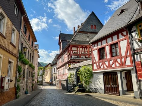 Kopfsteinpflasterstraße in Braubach gesäumt von bunten Fachwerkhäusern mit rot-weißen Fassaden unter blauem Himmel mit weißen Wolken.