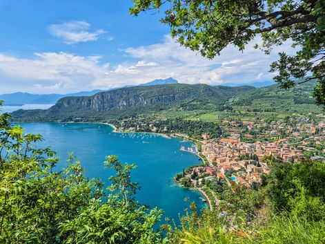 Panoramic view of a town along Lake Garda's turquoise bay, framed by green foliage. Mountains rise behind the settlement with boats dotting the water.