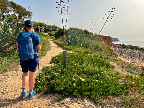 Wanderer mit blauem Rucksack auf sandigem Küstenweg der Rota Vicentina. Agaven und Meeresklippen unter bewölktem Himmel sichtbar.