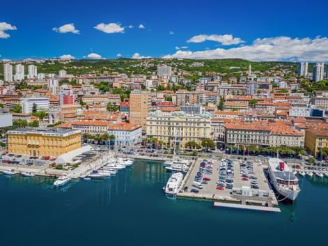 Aerial view of Rijeka harbor in Kvarner Bay with moored yachts, parking area, and historic yellow buildings along the waterfront under blue sky.