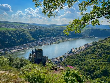 Aerial view of Burg Katz castle overlooking the Rhine River and St. Goarshausen town, surrounded by green hills under blue sky with clouds.