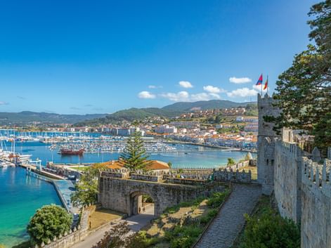 Blick von Festungsmauern in Baiona auf Yachthafen mit Segelbooten, Küstenstadt und grüne Hügel unter blauem Himmel.