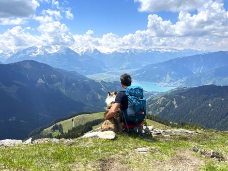 Wanderer mit Rucksack und Hund auf Berggipfel im Pinzgau mit Blick auf den türkisfarbenen Zeller See umgeben von Alpengipfeln.