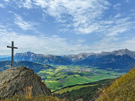 Summit cross on rocky peak overlooking green valley and Kitzbühel Alps mountain range under blue sky with scattered clouds.
