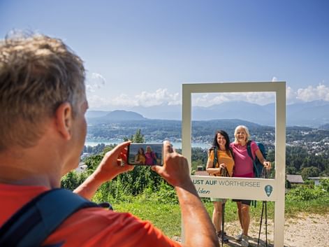Man taking photo of two women posing at Velden photo frame with lake and mountains in background. Clear blue sky over Carinthian landscape.