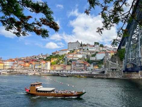 Traditional wooden boat on the Douro River with Porto's colorful hillside buildings and bridge visible. Blue sky with white clouds above the cityscape.