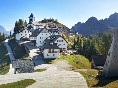 Monte Lussari mountain village with white church and traditional buildings on a hillside, surrounded by forests and rocky peaks under blue sky.