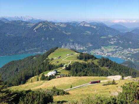 Panoramablick vom Schafberg mit grünen Almwiesen, gewundenen Wegen, türkisem See und Bergketten im Salzkammergut.