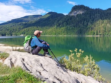 Female hiker with backpack and poles sitting on rock at Schwarzensee lake, Salzkammergut. Forested mountains reflect in calm green water.
