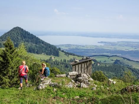 Zwei Wanderer mit Rucksäcken auf einem Bergweg mit Blick auf den Chiemsee. Eine Holzhütte steht auf Felsen, bewaldete Gipfel und Tal darunter.