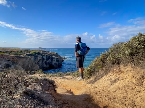 Wanderer mit blauem Rucksack auf sandigem Weg mit Blick auf felsige Klippen und blauen Atlantik bei Porto Covo an der Rota Vicentina.
