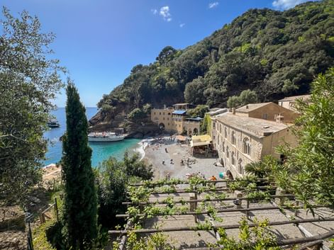 San Fruttuoso bay with historic abbey buildings, pebble beach, and turquoise water. Lush green hillside rises behind the beige stone structures.