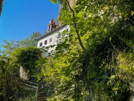 White hermitage building with bell tower perched on steep rocky cliff surrounded by lush green trees under blue sky in Salzburg region.