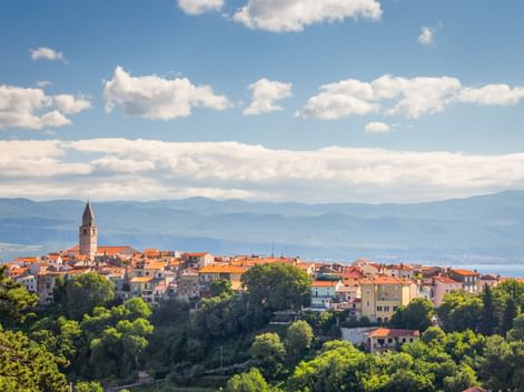 Coastal town of Vrbnik on island Krk with church tower, terracotta roofs, and lush greenery. Mountains and blue sea visible in background.