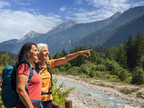 Two women hikers with backpacks admiring mountain view near Villach. One points across a turquoise river toward forested Alpine peaks.