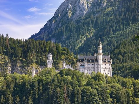 Neuschwanstein Castle nestled among dense forests with dramatic rocky mountain peaks rising behind it under a blue sky.