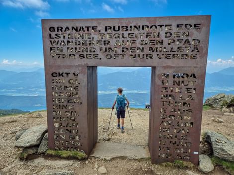 Wanderer mit Trekkingstöcken geht durch das metallene Granattor mit ausgestanztem Text auf einem Berggipfel. Bergketten im Hintergrund unter blauem Himmel.