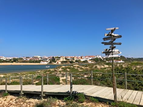 Wooden boardwalk with directional signpost on coastal path overlooking river and Vila Nova de Milfontes town under clear blue sky.