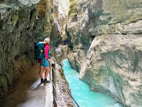 Wanderin mit Rucksack auf Holzsteg durch die Partnachklamm mit türkisfarbenem Wasser zwischen Kalksteinwänden.