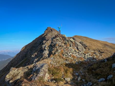 Female hiker with raised arms celebrating on rocky mountain summit in the Kitzbühel Alps under clear blue sky with mountain ranges visible.