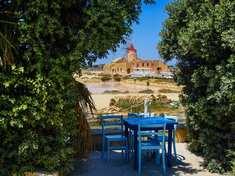 Blick auf Trapani Windmühle durch Gartenterrasse Blaues Outdoor-Essset auf Terrasse umrahmt von grünem Laub, mit Blick auf historische Windmühle und Salzfelder von Trapani, Sizilien.