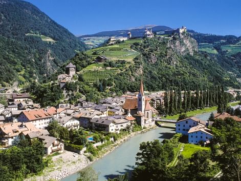 Alpine town of Klausen with church spire, river, and historic castles on hillsides. Mountains and vineyards visible in background under blue sky.