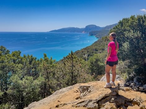 Woman in pink shirt standing on rocky viewpoint overlooking turquoise Kvarner Bay with green hills and distant mountains under blue sky.