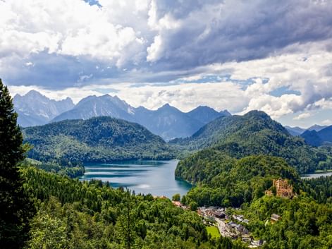Aerial view of Hohenschwangau village with yellow castle tower, surrounded by forested hills and blue Alpine lakes, with mountain peaks in background.