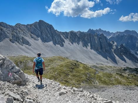 Hiker with backpack walking on rocky mountain path in Karwendel with dramatic jagged peaks and scree slopes under blue sky with clouds.