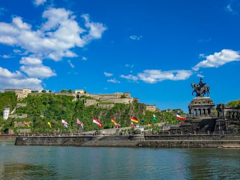 Deutsches Eck Denkmal mit Reiterstatue in Koblenz, bunte Flaggen am Rheinufer, Festung Ehrenbreitstein auf dem Hügel.
