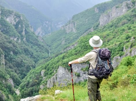 Hiker with backpack and walking stick overlooking the lush green Rakitnica gorge in Bosnia, surrounded by steep limestone cliffs and forest.