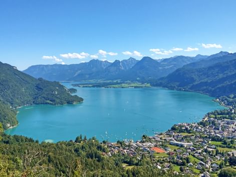 Aerial view of turquoise Wolfgangsee surrounded by mountains and green forests. A lakeside town is visible on the right shore under clear blue sky.