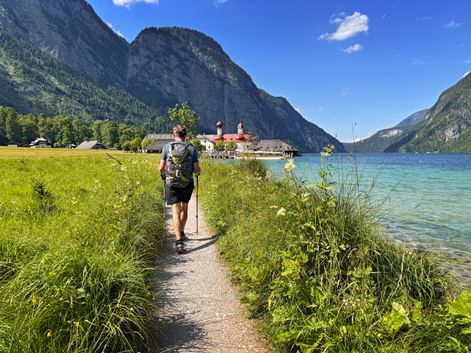 Hiker with backpack walking on path beside turquoise Königssee lake toward St. Bartholomä church with red onion domes, surrounded by Alps.