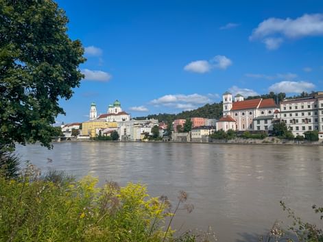 View of Passau's historic old town across the Danube River, showing baroque churches with green domes and colorful buildings under blue sky.