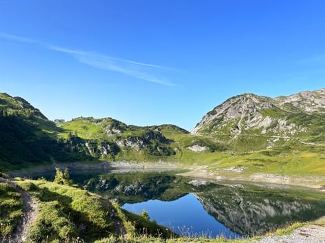 Bergsee Formarinsee spiegelt Felsgipfel unter blauem Himmel. Grüne Almwiesen und Wanderweg entlang des Ufers am Lechweg sichtbar.