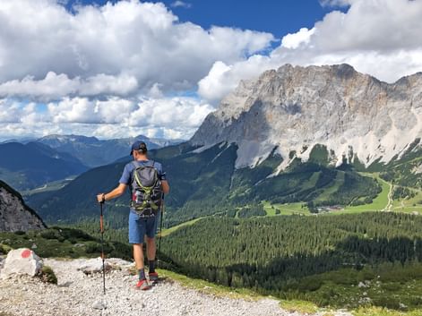 Wanderer mit Rucksack und Wanderstöcken auf felsigem Weg, mit Blick auf dramatisches Alpental mit Zugspitze-Gebirge und grünen Wäldern.