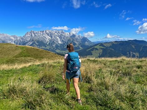 Female hiker with blue backpack walking on grassy alpine meadow with dramatic mountain peaks in background under blue sky with clouds.