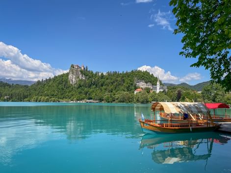 Traditional wooden boat on turquoise Lake Bled with Bled Castle on a forested cliff and a white church tower visible on the shore.