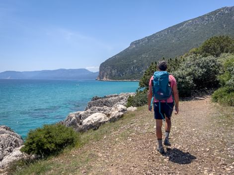 Hiker with backpack walking on rocky coastal path in Northern Dalmatia. Turquoise Adriatic Sea and green mountain slope visible.